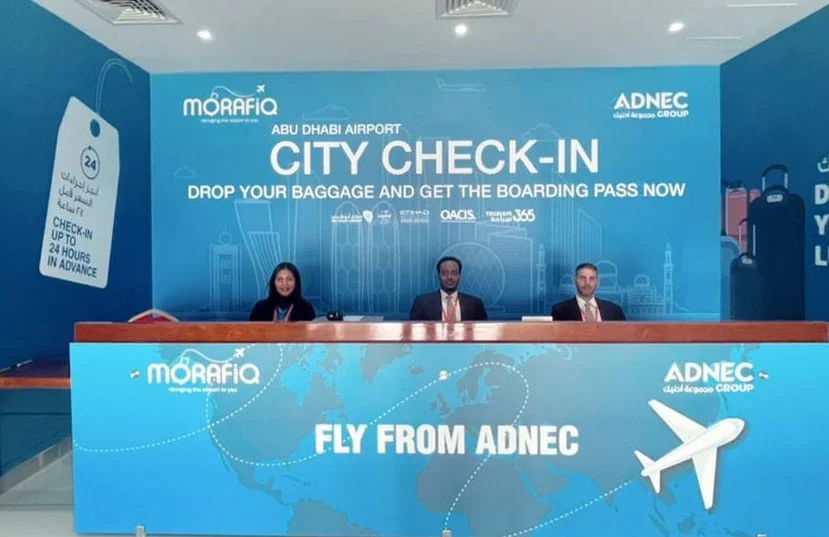 Three staff members sit behind a check-in counter for Abu Dhabi Airport's City Check-In service, which allows passengers to drop their baggage and receive boarding passes. The backdrop includes travel-related graphics and informational text about the service.