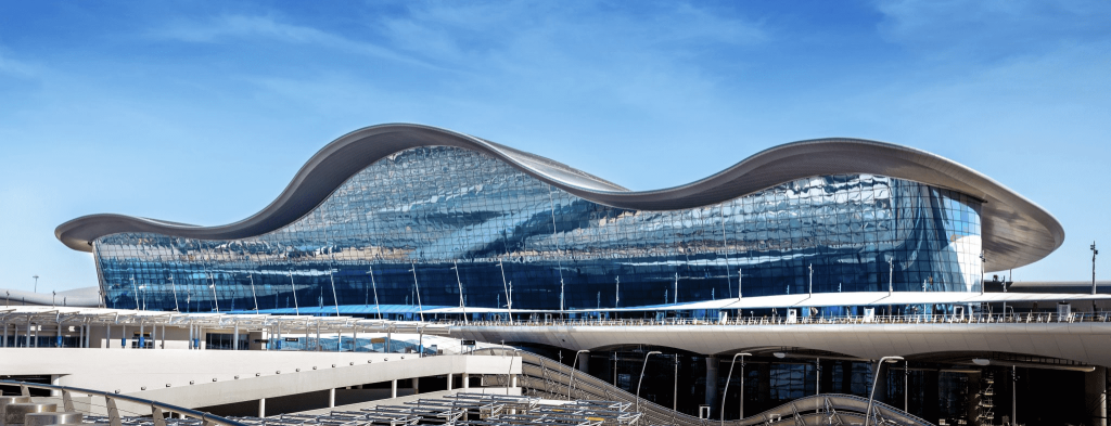 A modern airport terminal with a distinctive wave-like roof and a glass facade reflecting the blue sky. The architectural design features curving lines and sleek surfaces, with a network of pathways and platforms in the foreground.