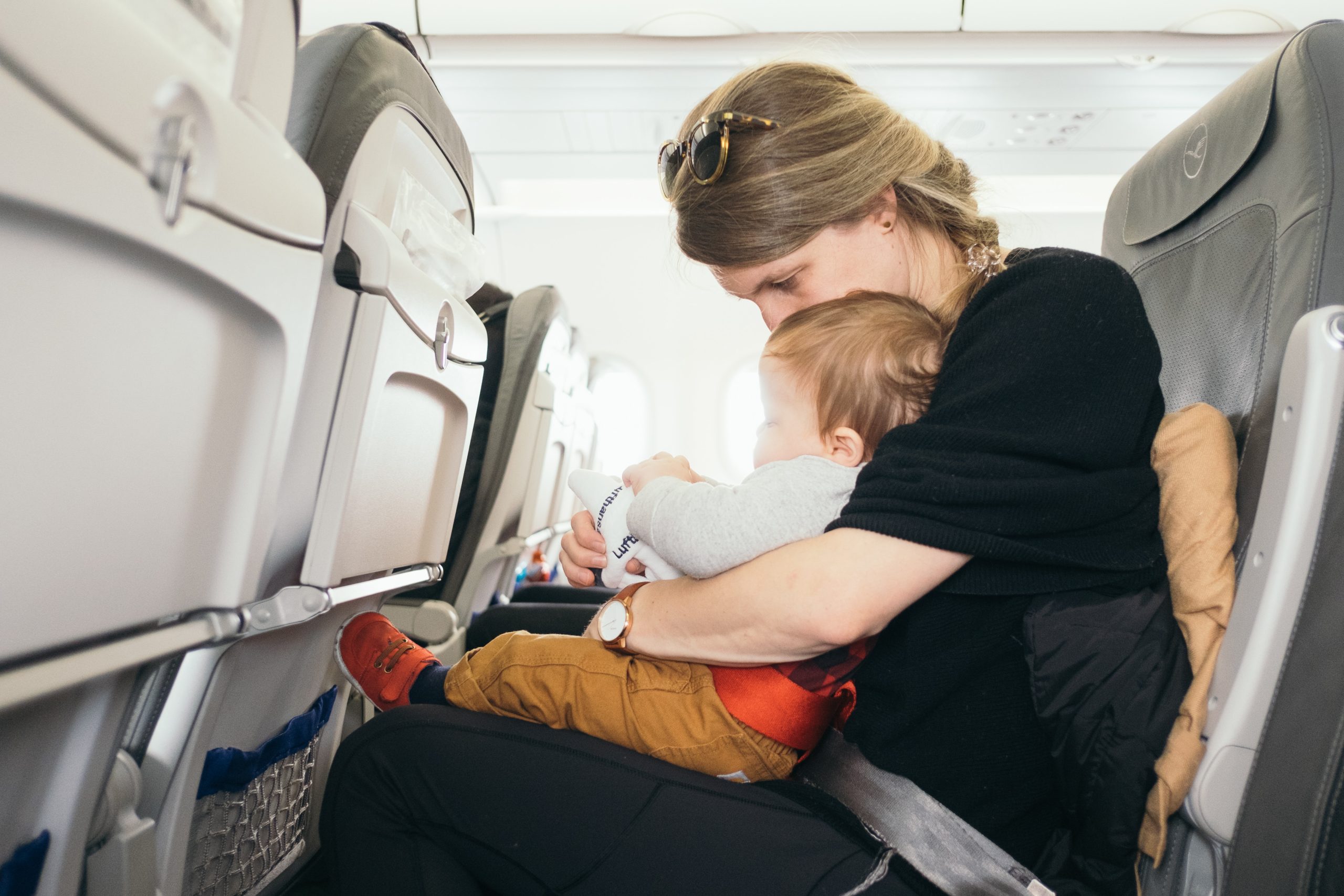 A woman with glasses on her head sits in an airplane seat, holding a baby on her lap. The baby is focused on something in their hands. Both are fastened with a seatbelt. The airplane's interior and rows of seats are visible.