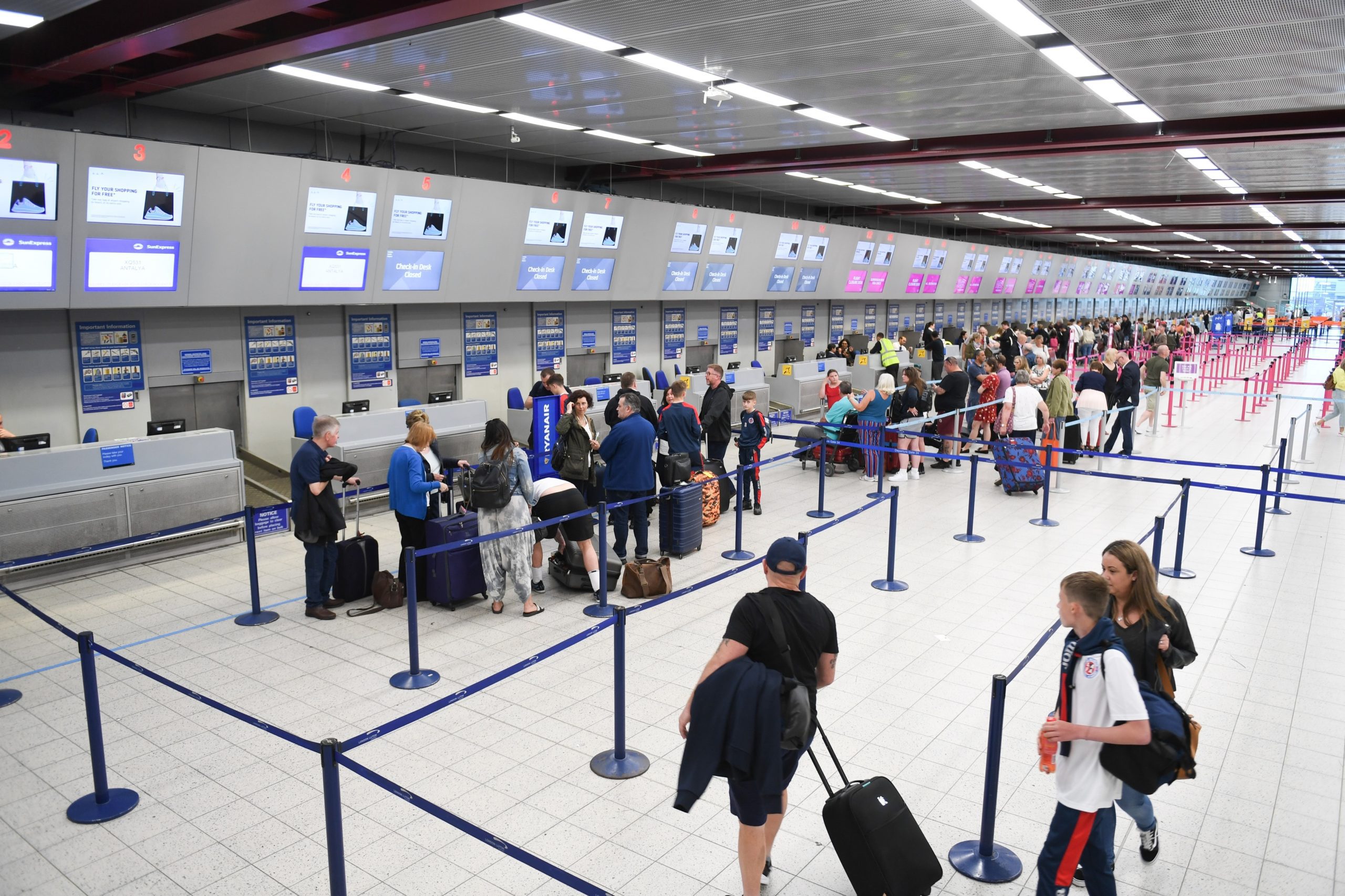 A busy airport check-in area with multiple counters and digital screens. Passengers are standing in line, managing luggage, and waiting to check in. Blue stanchions organize the queues, and staff are assisting travelers. The area is brightly lit.