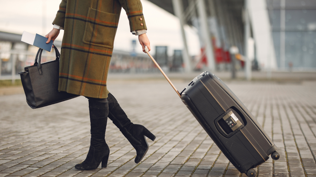 A Person in a green coat and black boots walking on a cobblestone surface while pulling a black rolling suitcase with one hand and holding a black handbag and a passport in the other. The background features an industrial-style building.