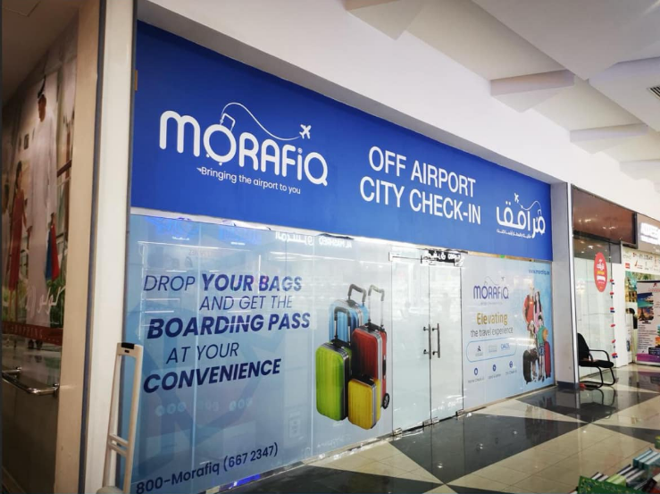 A storefront with blue signage reading "MORAFIQ" and "OFF AIRPORT CITY CHECK-IN." The storefront has images of luggage and text promoting services like bag drop-off and boarding pass collection. Contact information and the slogan "Bringing the airport to you" are also visible.
